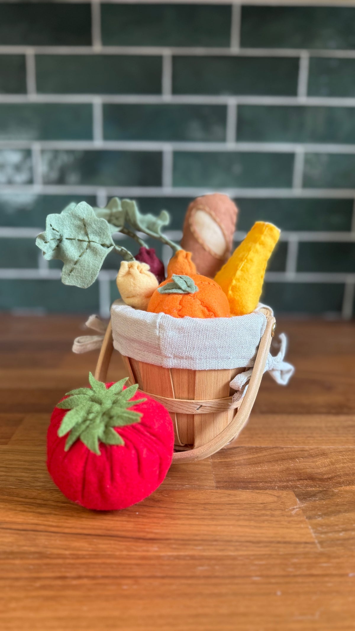 A felt play kitchen basket filled with fabric vegetables and fruits, placed on a wooden table in front of a green subway tile wall. A red felt tomato sits in the foreground.